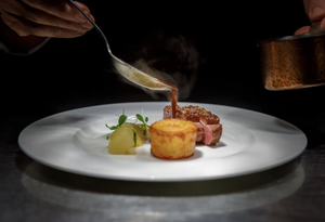 Sauce being poured over a plated meat dish with vegetables and a golden potato garnish