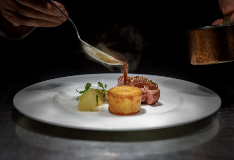 Sauce being poured over a plated meat dish with vegetables and a golden potato garnish